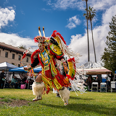 Indigenous man in headdress and decorative clothing and boots dancing on SCU's campus during the annual Powwow