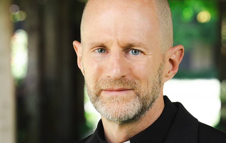 Man with bald head and beard, wearing black shirt and religious collar, standing in walkway next to Mission church. 