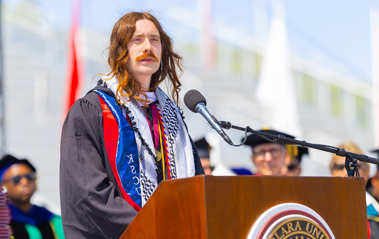 Simon Lanzoni (long hair and mustache) wears graduation gown, stands at podium during commencement. 
