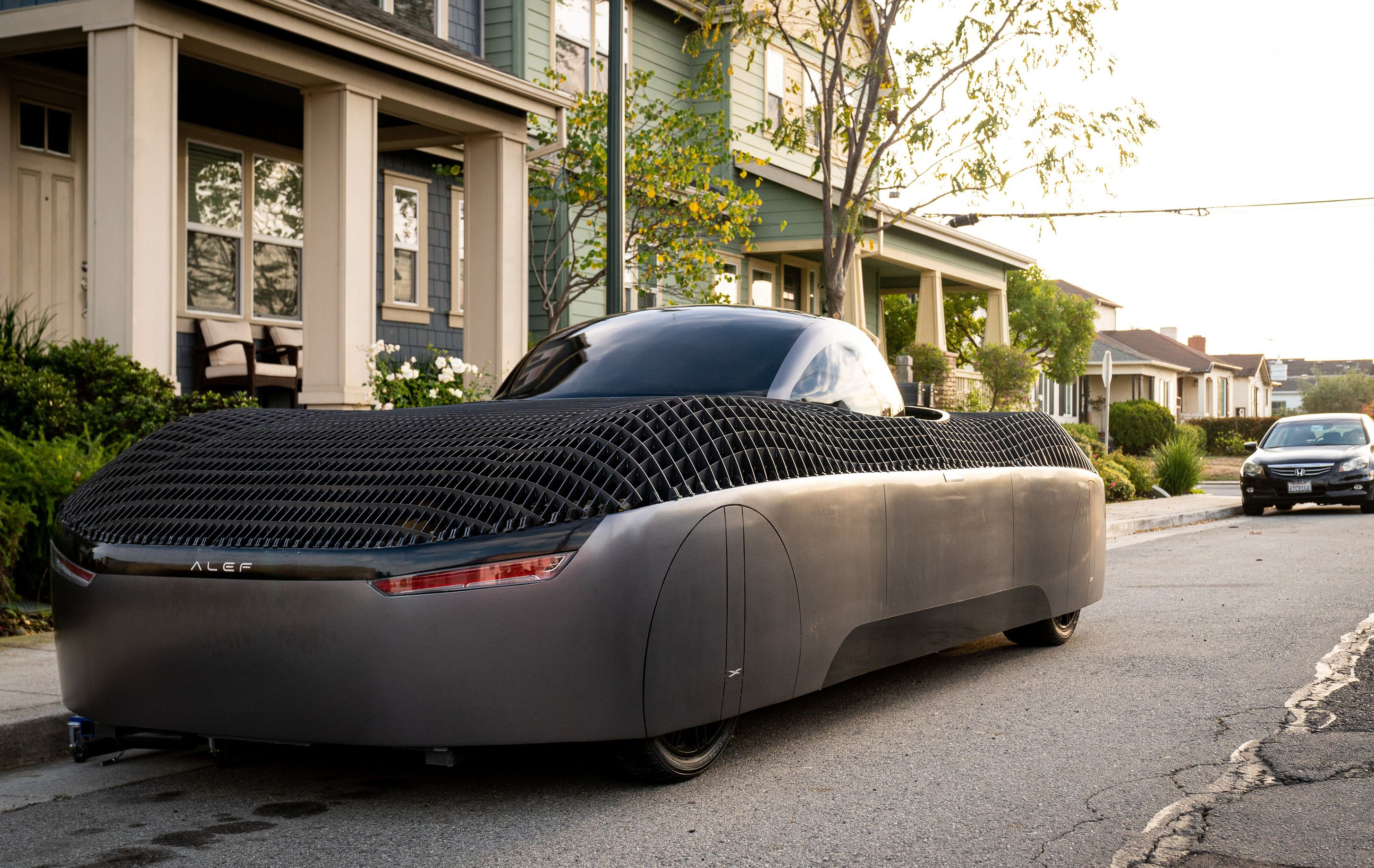 A futuristic looking flying car parked on the street outside of a residential home. 