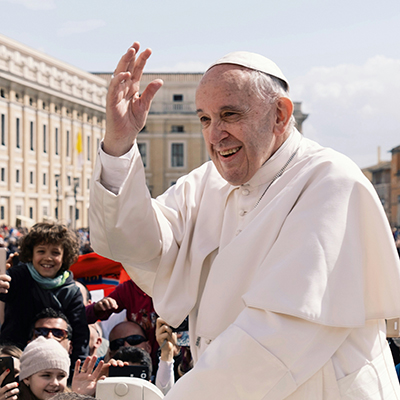 Pope Francis raising his hand as he addresses people with a smile